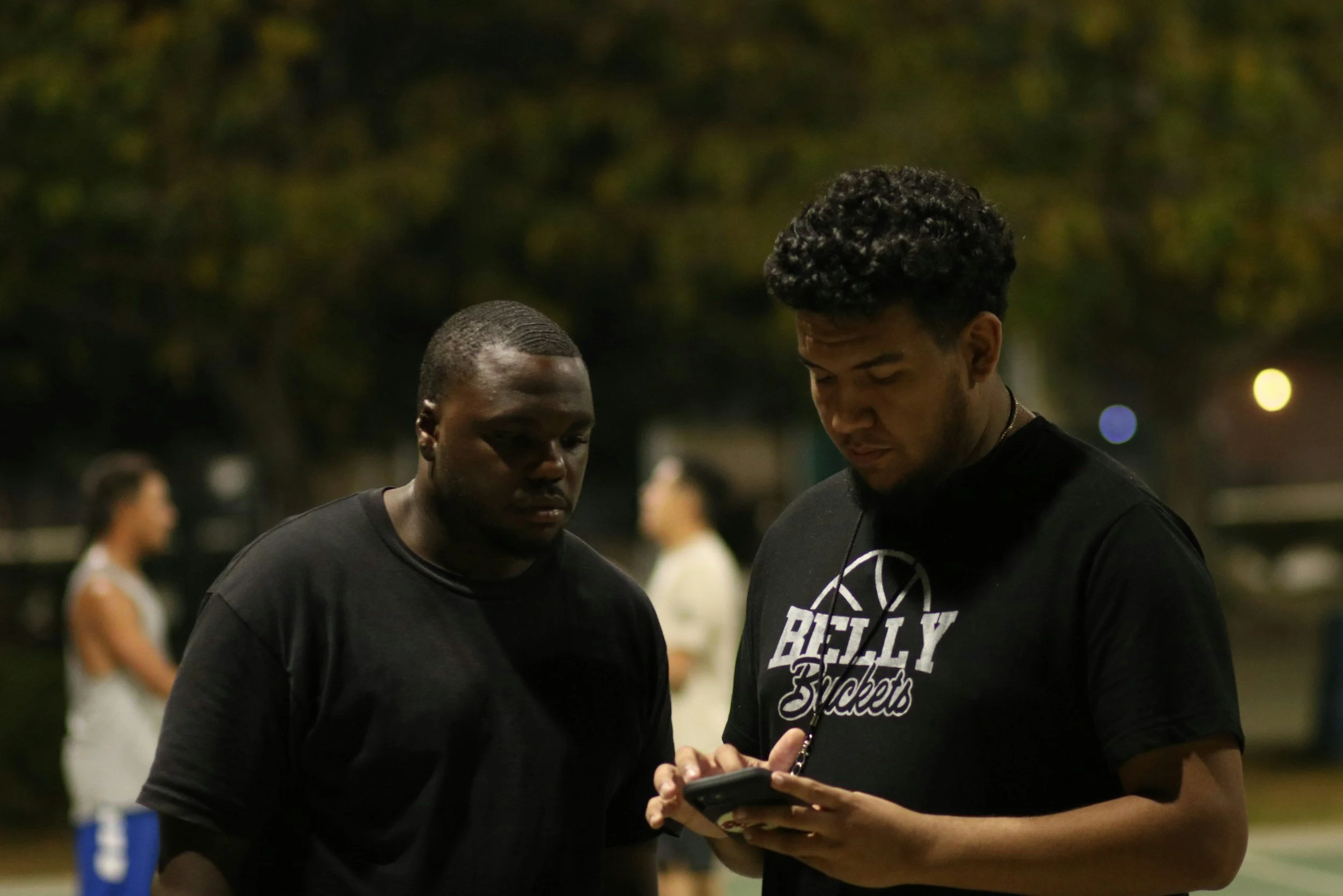 Two young men standing close together outdoors at night, looking at a smartphone held by one of them.