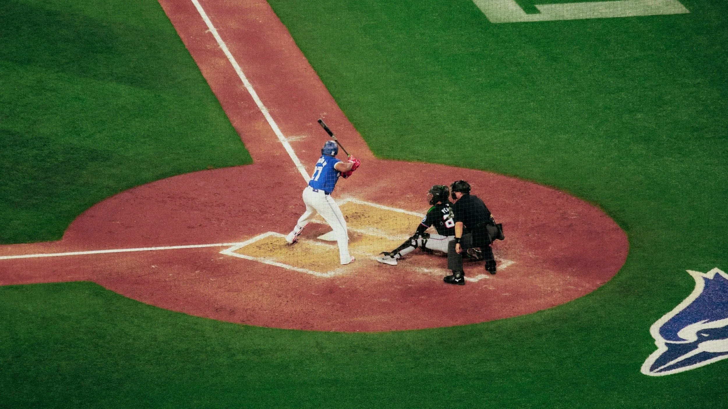 A baseball player in a blue jersey and white pants at home plate prepared to swing, with umpires nearby, on a baseball field with green grass and part of a logo visible on the turf.