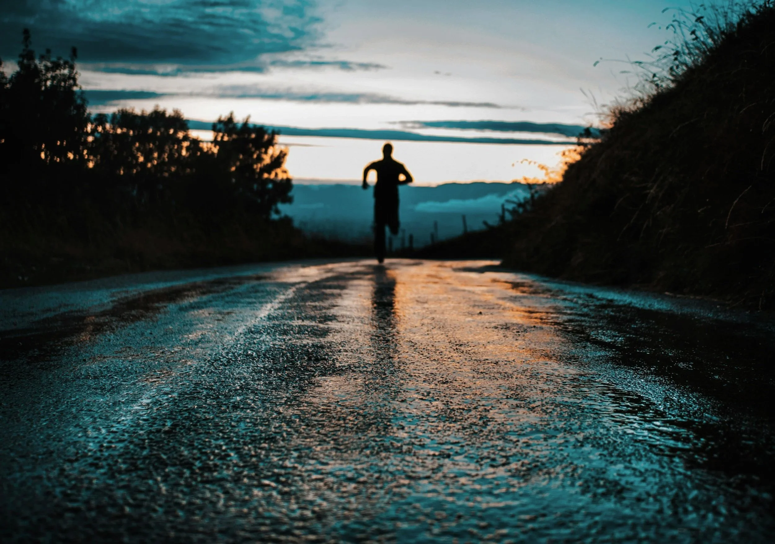 A person running on a wet road at sunset or sunrise, with the sky showing dark clouds and a hint of orange light in the background.