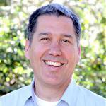A smiling man with short dark hair wearing a light blue collared shirt outdoors with trees and sunlight in the background.