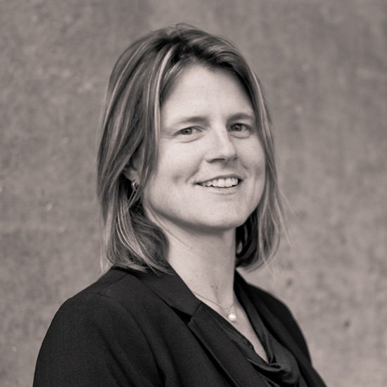 A woman with shoulder-length hair, smiling, wearing a dark blazer and pearl necklace, standing against a neutral background.