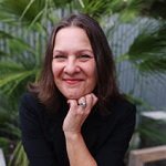 A woman with shoulder-length brown hair smiling and resting her chin on her hand, outdoors with green foliage in the background.