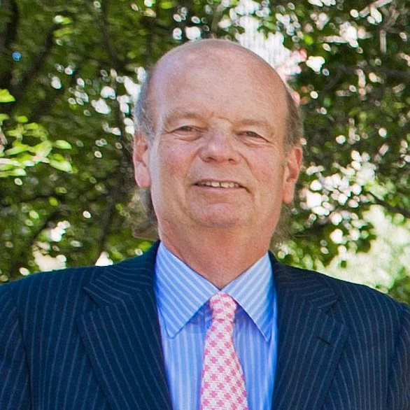 A middle-aged man with a receding hairline wearing a dark pinstripe suit, blue and white striped shirt, and pink patterned tie. He is smiling and standing outdoors with green foliage in the background.