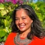A woman smiling outdoors with a background of trees and blue sky.
