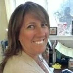 A woman with shoulder-length brown hair smiling in an office setting with windows and computer monitors in the background.