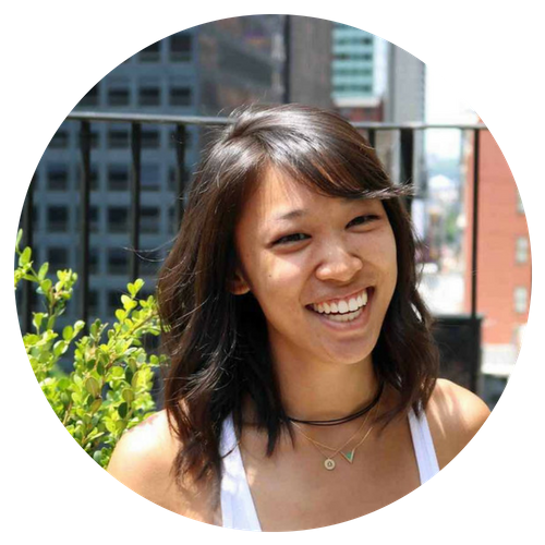 A young woman smiling outdoors in an urban setting with buildings and a railing in the background.