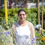 A woman standing outdoors in a garden with yellow posts, wearing sunglasses, a black headscarf, a beige cardigan, and a white shirt, surrounded by purple flowers and green plants.