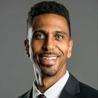 Professional headshot of a smiling man with short curly hair, wearing a dark suit and white shirt against a gray background.