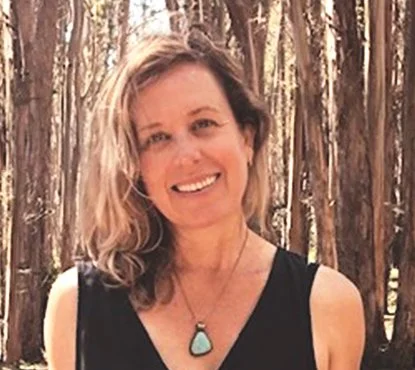 A picture of Jenn Weiss, coach and consultant, with wavy blonde hair smiling outdoors in front of trees, wearing a black sleeveless top and a turquoise pendant necklace.
