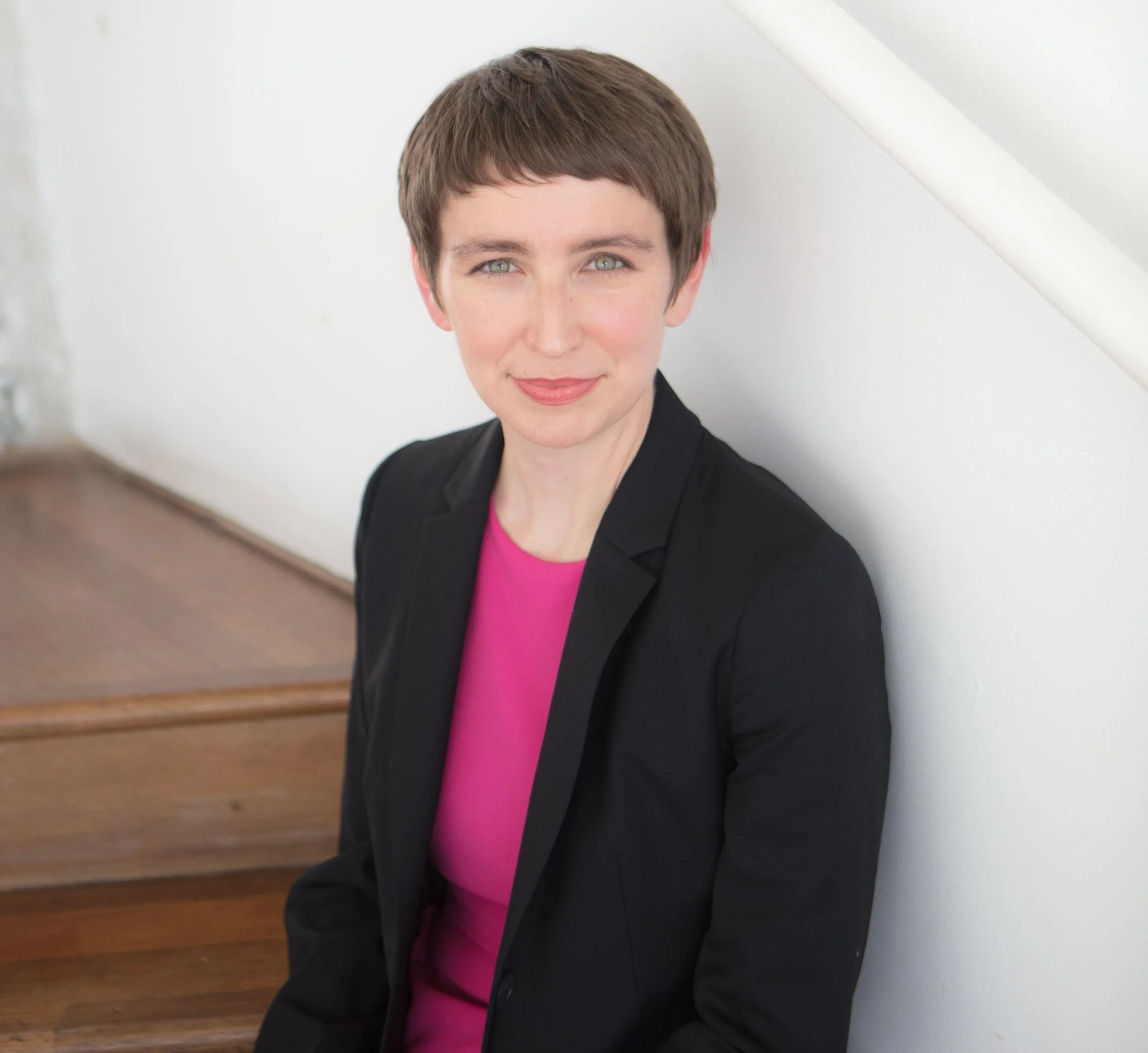 A woman with short brown hair, light skin, and blue eyes, wearing a black blazer and pink shirt, sitting on a wooden staircase with a white wall background.