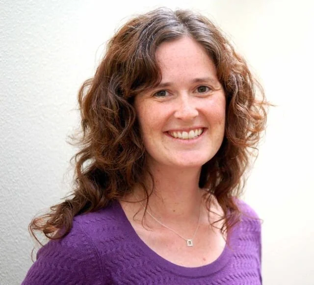 A woman with shoulder-length curly brown hair wearing a purple sweater and a silver necklace, smiling against a plain light-colored background.