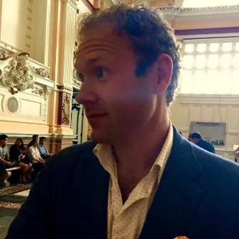 A man with curly hair wearing a suit and white shirt sitting in a large, ornate room with high ceilings, tall windows, and decorative woodwork.
