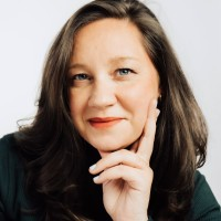 A woman with long brown hair and blue eyes, resting her face on her hand and smiling softly, wearing a dark top.
