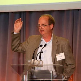 A man wearing glasses and a suit jacket speaking at a podium during a conference.