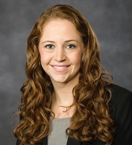 A young woman with long, curly red hair, smiling, wearing a black blazer and gray top, against a gray background.