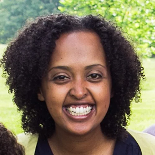 A woman with curly black hair smiling outdoors with a blurred green background.
