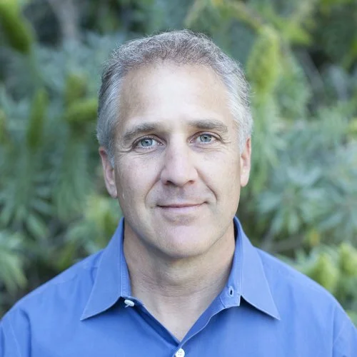 Portrait of a middle-aged man with gray hair wearing a blue collared shirt, standing outdoors with green foliage in the background.