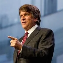 A man with medium-length brown hair, wearing a dark suit and red tie, is pointing with his right hand while speaking.