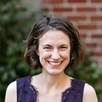 A woman with short, wavy brown hair, smiling outdoors in front of a brick wall and greenery.