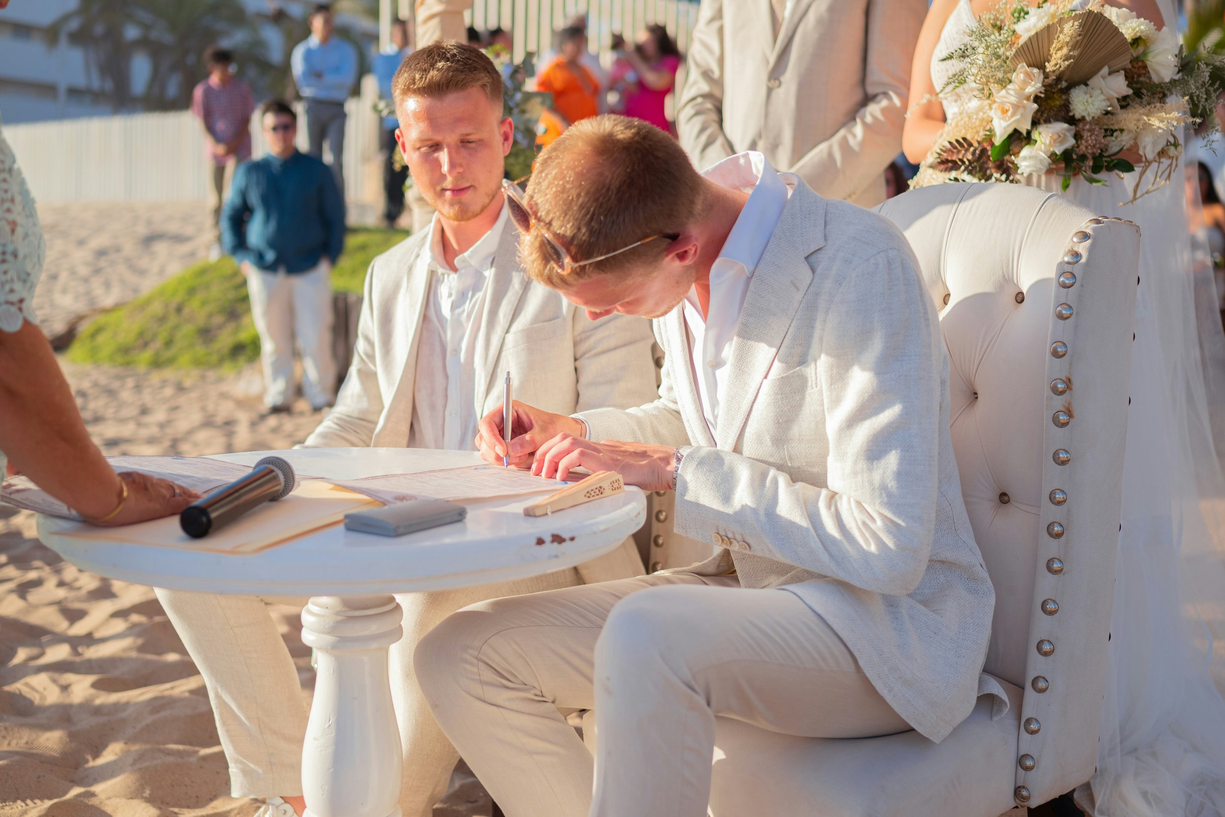 Twee mannen tekenen een huwelijksakte tijdens een bruiloft op het strand, omringd door gasten.