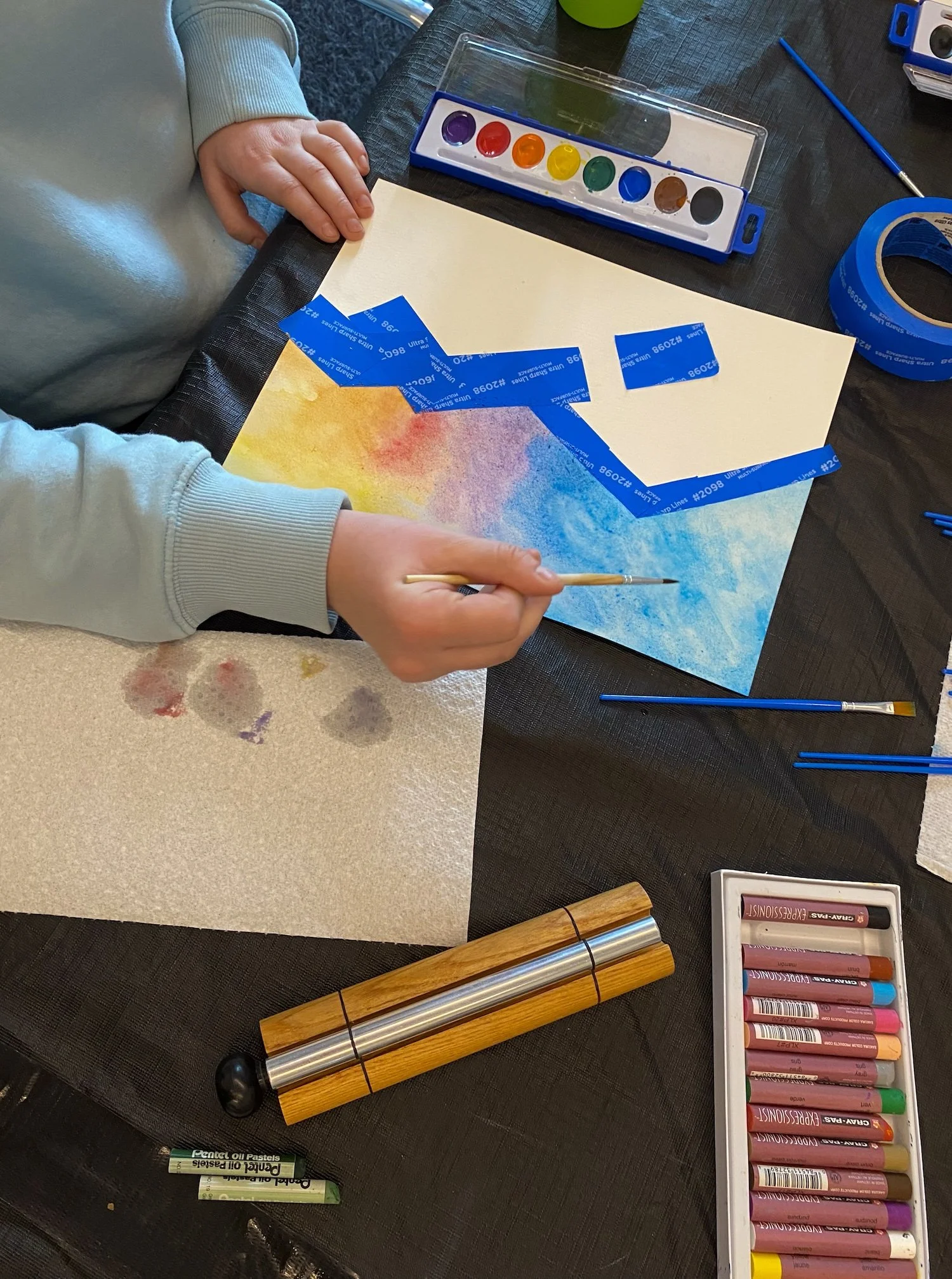 Person creating watercolor art with masking tape, paints, brushes, and pastels on a table.