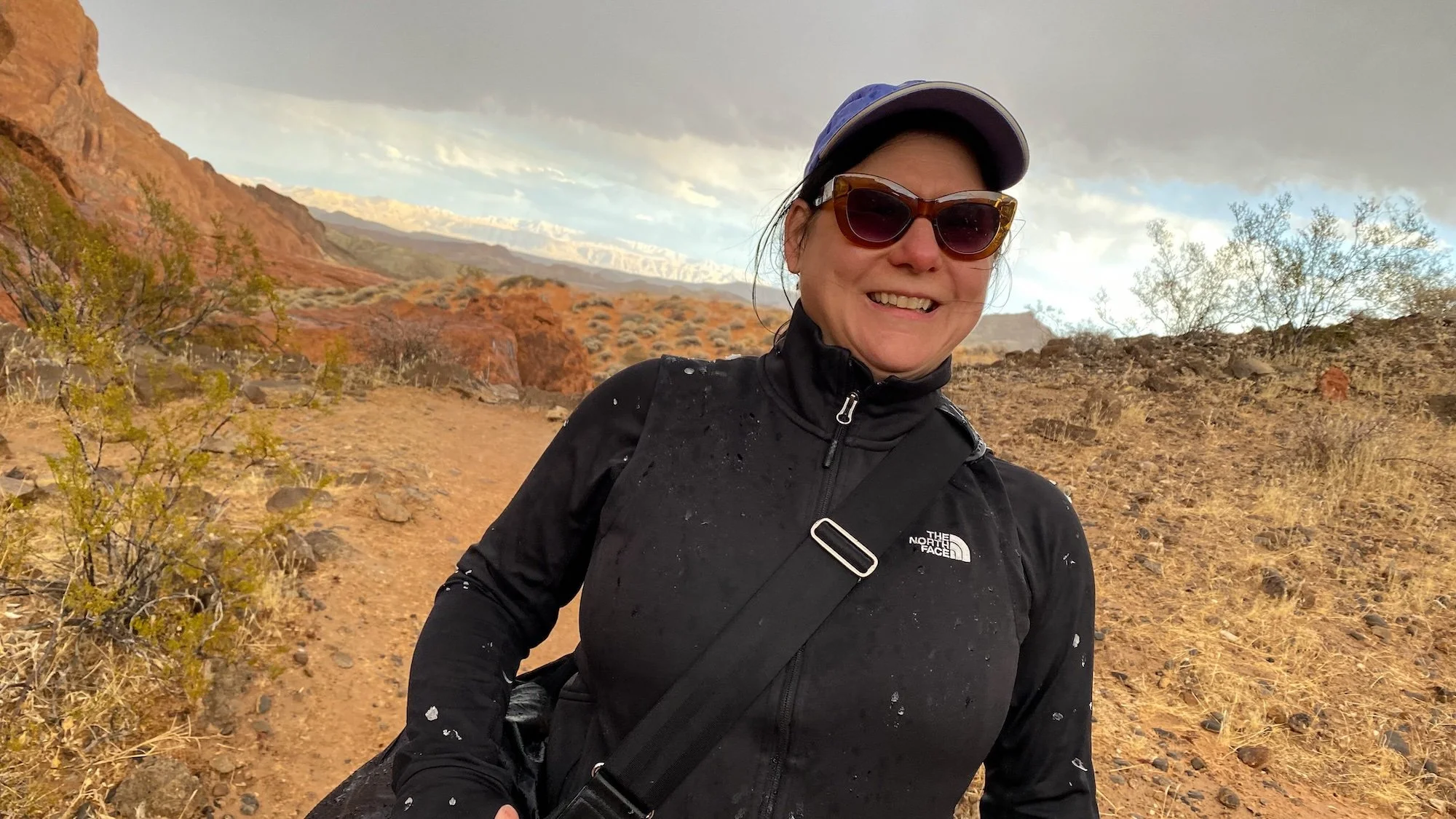 Therapist Dana Keeton smiling, wearing sunglasses and a black jacket walking on a dirt trail in a desert landscape with mountains and cloudy sky in the background.