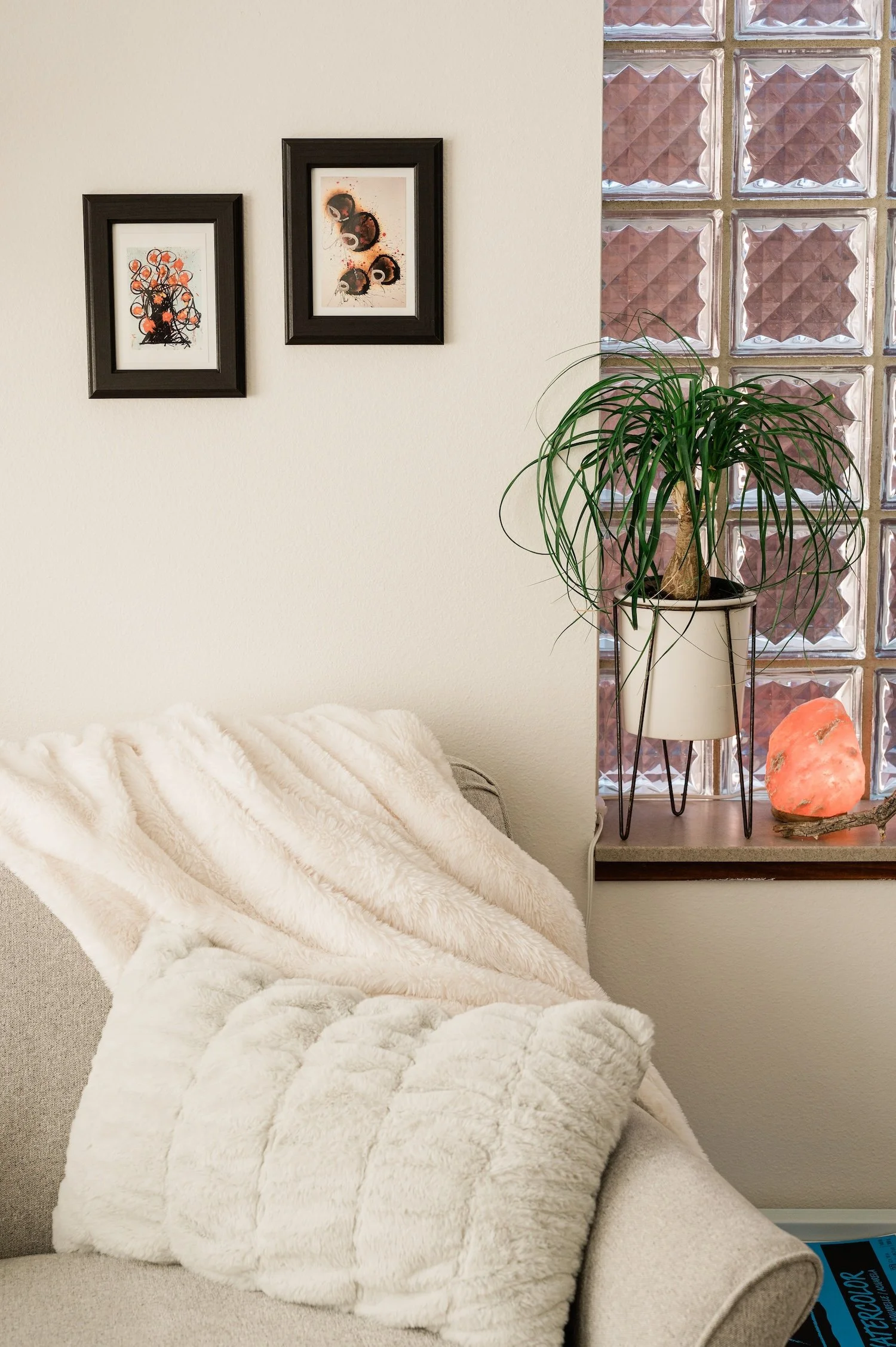 Dana Keeton's cozy therapy room in downtown Iowa City with a beige sofa, a plush white blanket, two framed art pictures on the wall, a potted plant on a window sill, and a Himalayan salt lamp.