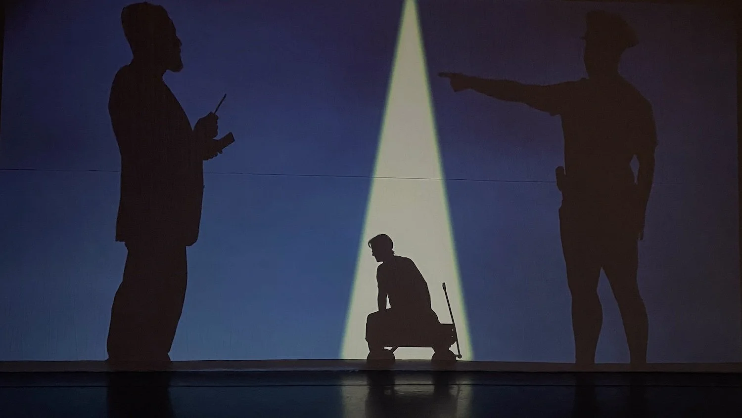 Silhouettes of three people, one of whom is seated on a small cart, with a spotlight shining on them against a blue background.