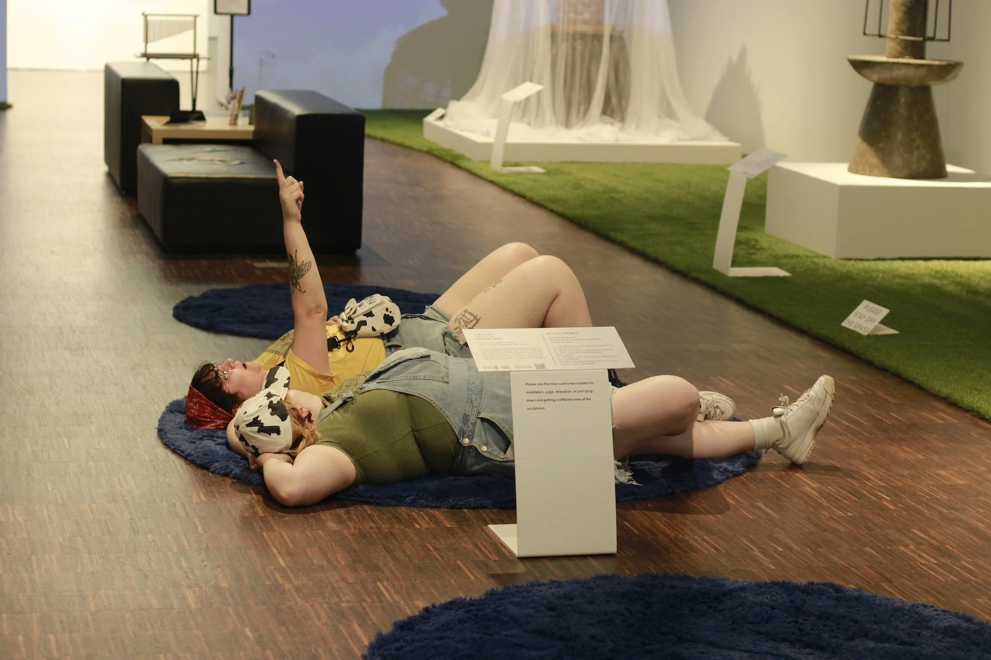 Two people lying on a blue rug on the floor in an art gallery among sculptures. One of the people is pointing at the ceiling
