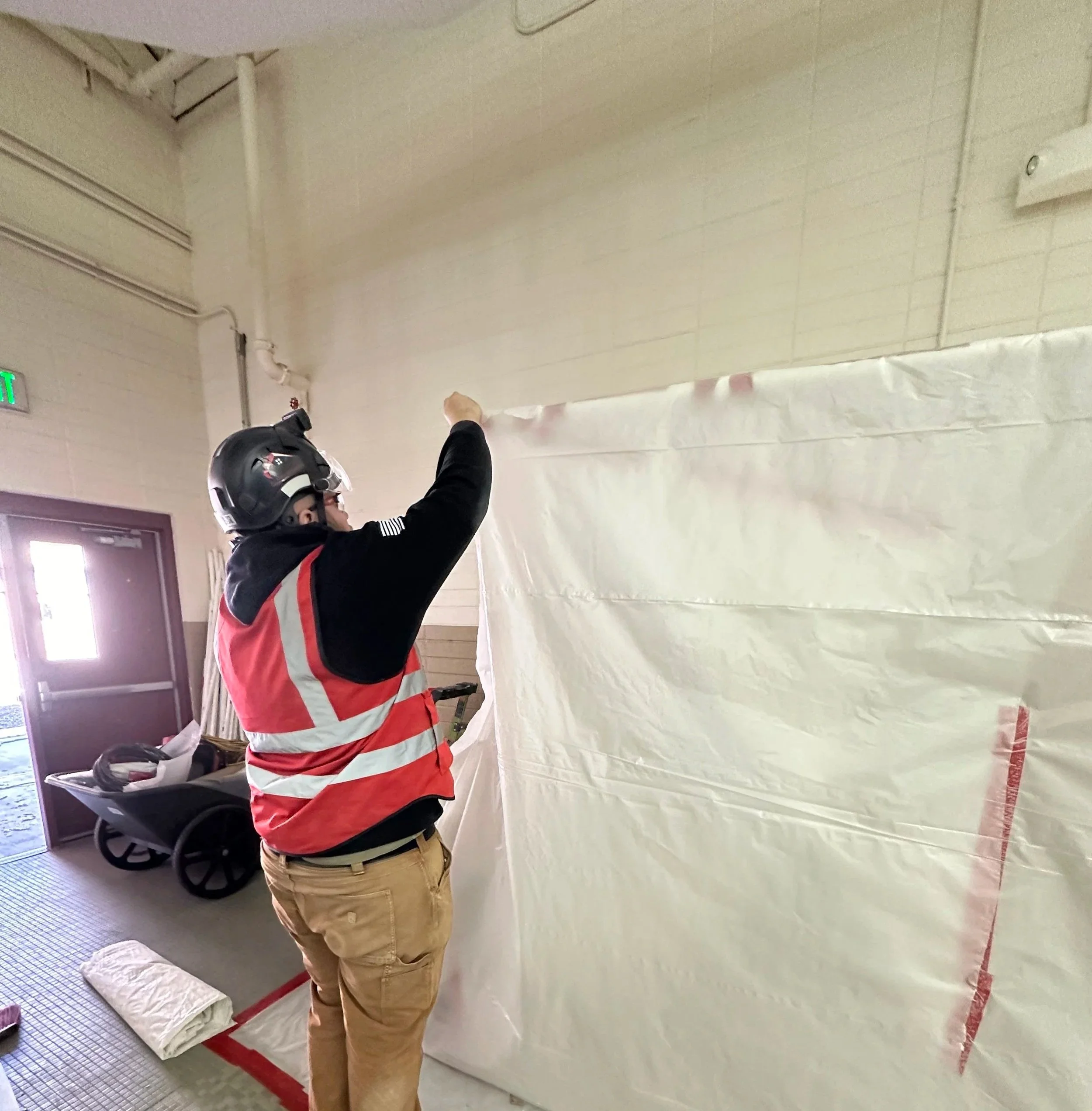 A person wearing a black helmet, a black long-sleeve shirt, and a red safety vest is drawing or writing on a large piece of white paper or plastic attached to the wall inside a building. There is a wheelbarrow and some rolled-up material on the floor nearby.