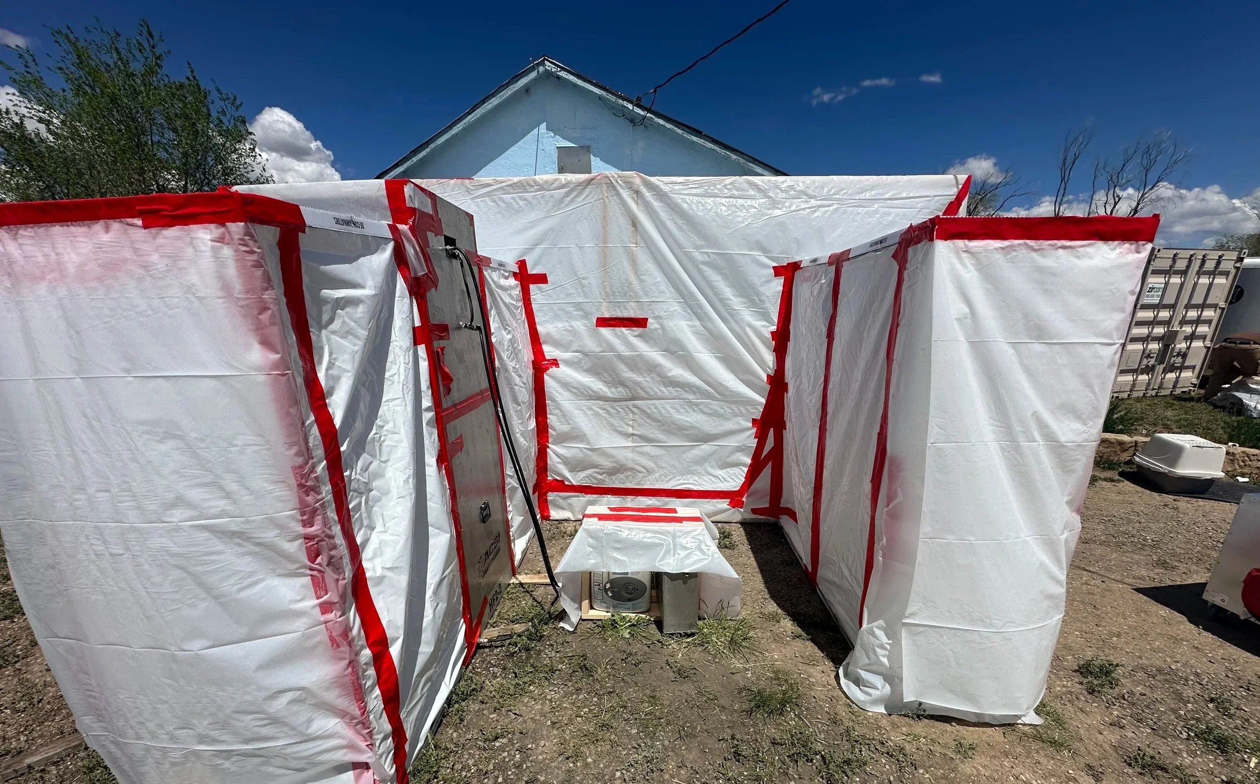 Temporary outdoor medical or testing tent with white fabric and red tape, set up in a yard with a small table and a house in the background.
