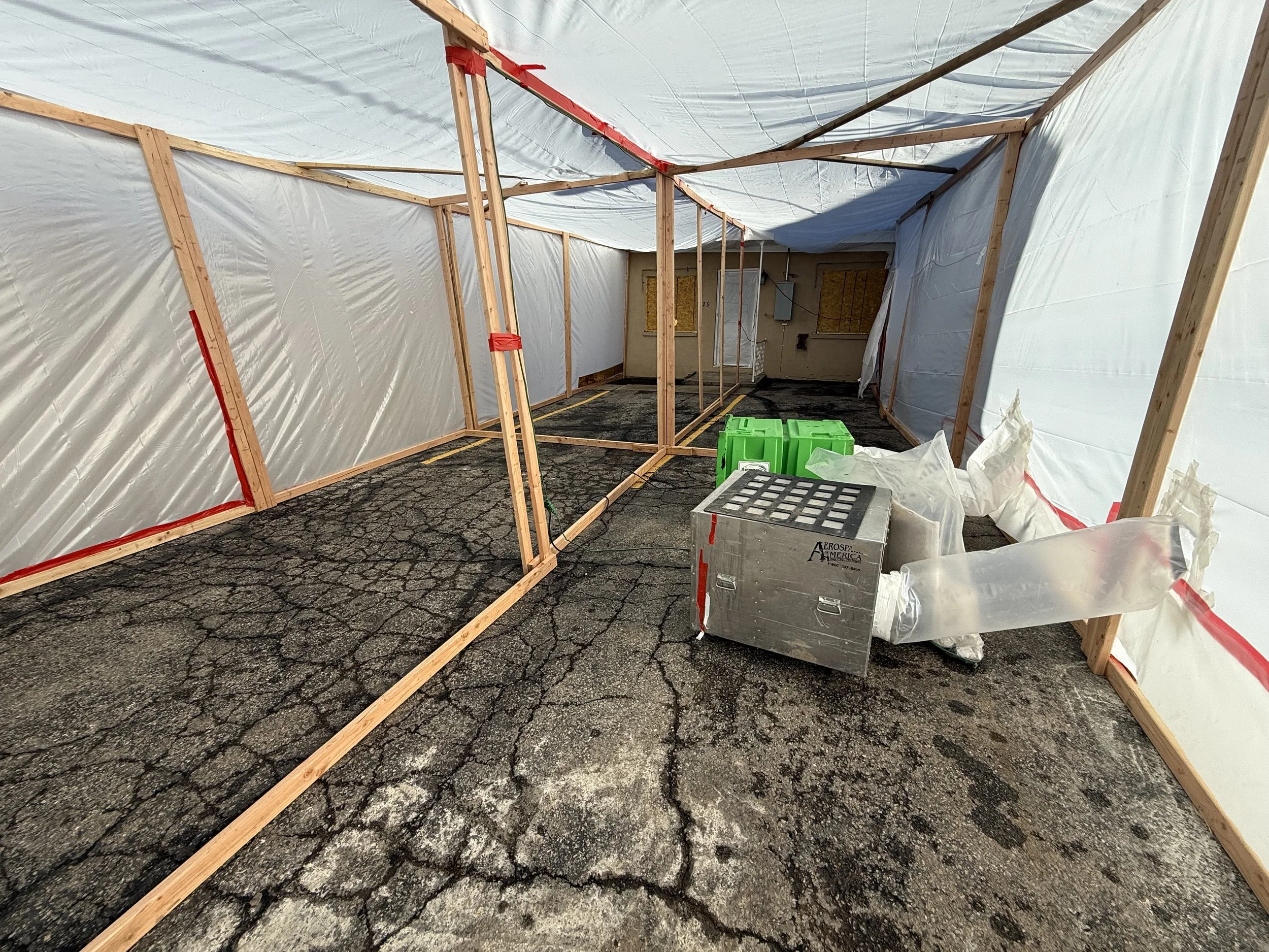 Construction site inside with wooden framing and white plastic sheeting walls, cracked asphalt floor, and various tools and equipment including a metal box, green containers, and wrapped objects.