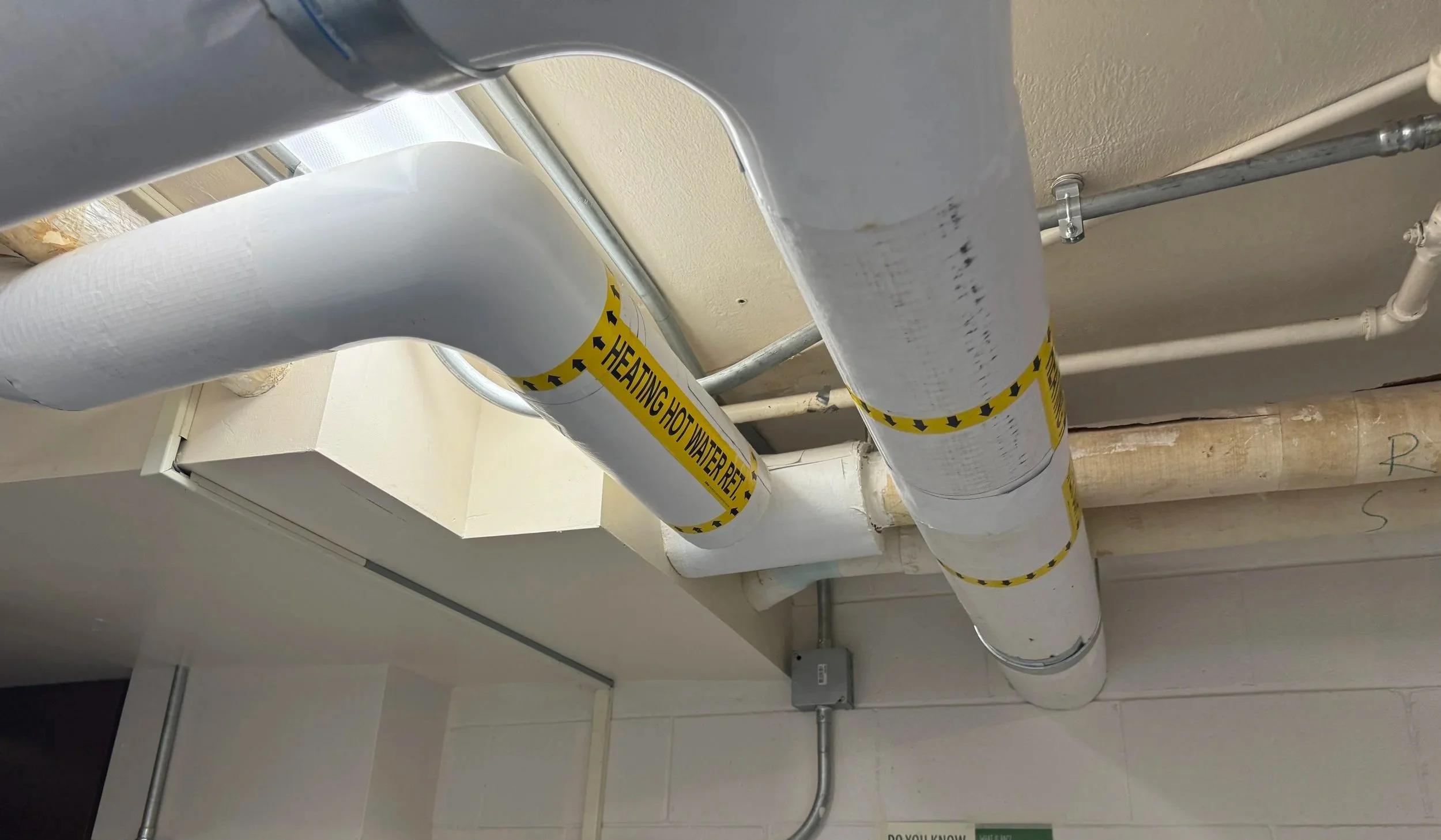Pipes in ceiling with yellow warning tape that reads 'Heating Hot Water Return'.