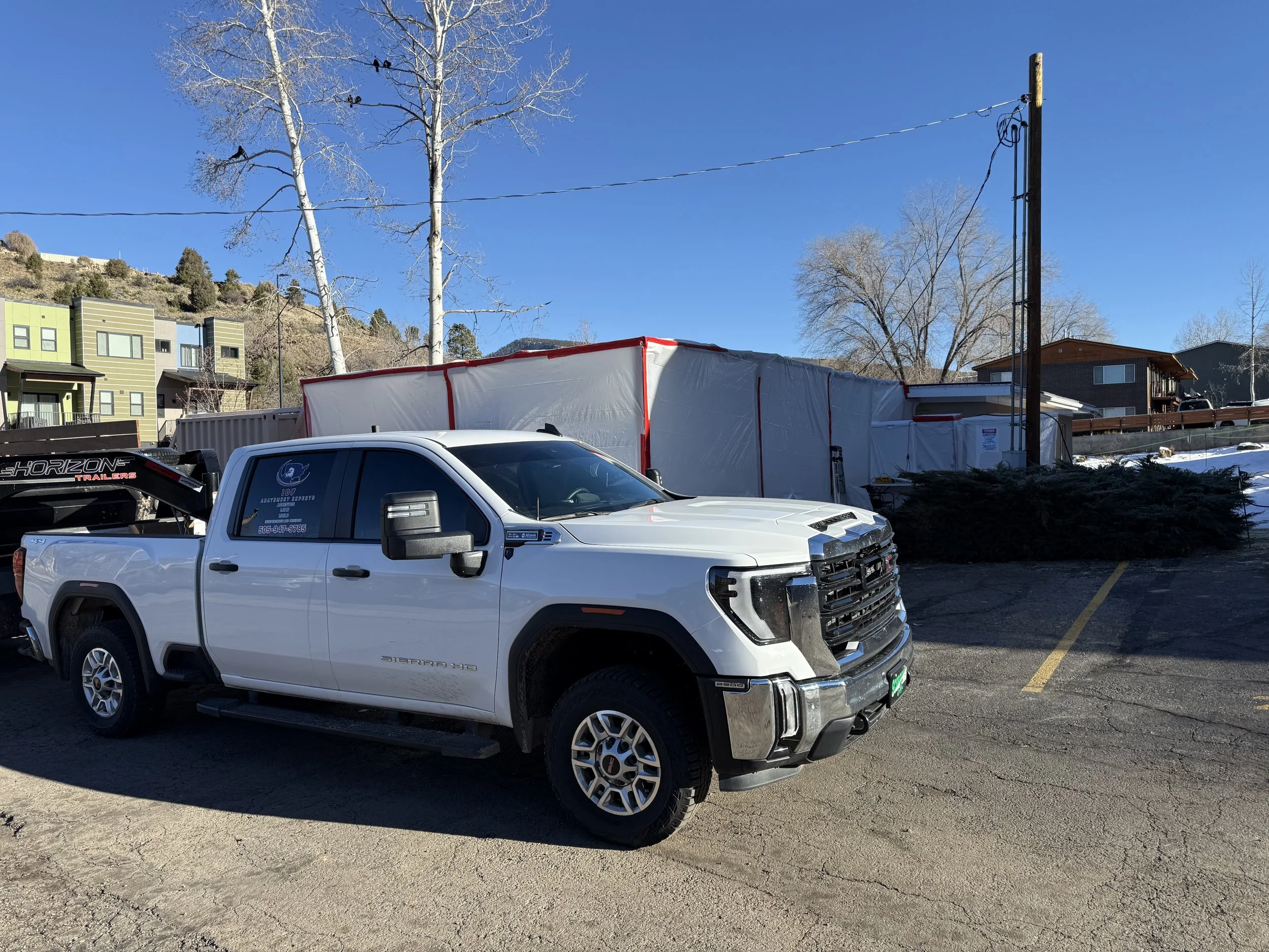 A white pickup truck parked next to a tall white and red-covered structure in a parking lot under a clear blue sky, with leafless trees and residential buildings in the background.