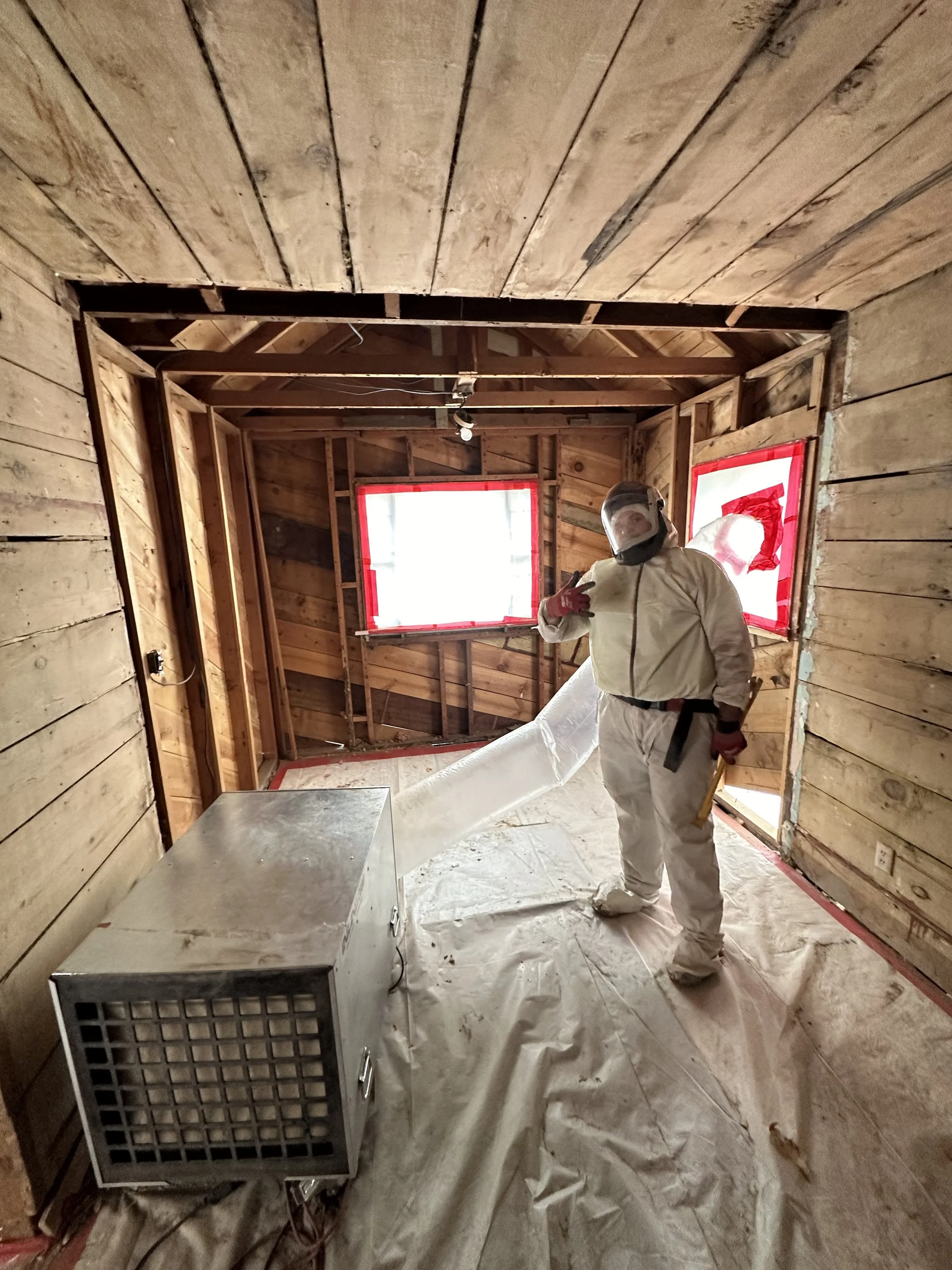 A person dressed in protective gear standing in a partially constructed room with wooden walls. The person is giving a thumbs-up gesture and is wearing a face shield. There is a small heater on the floor, and plastic sheeting covers the floor. The room has two small windows with red tape around them.