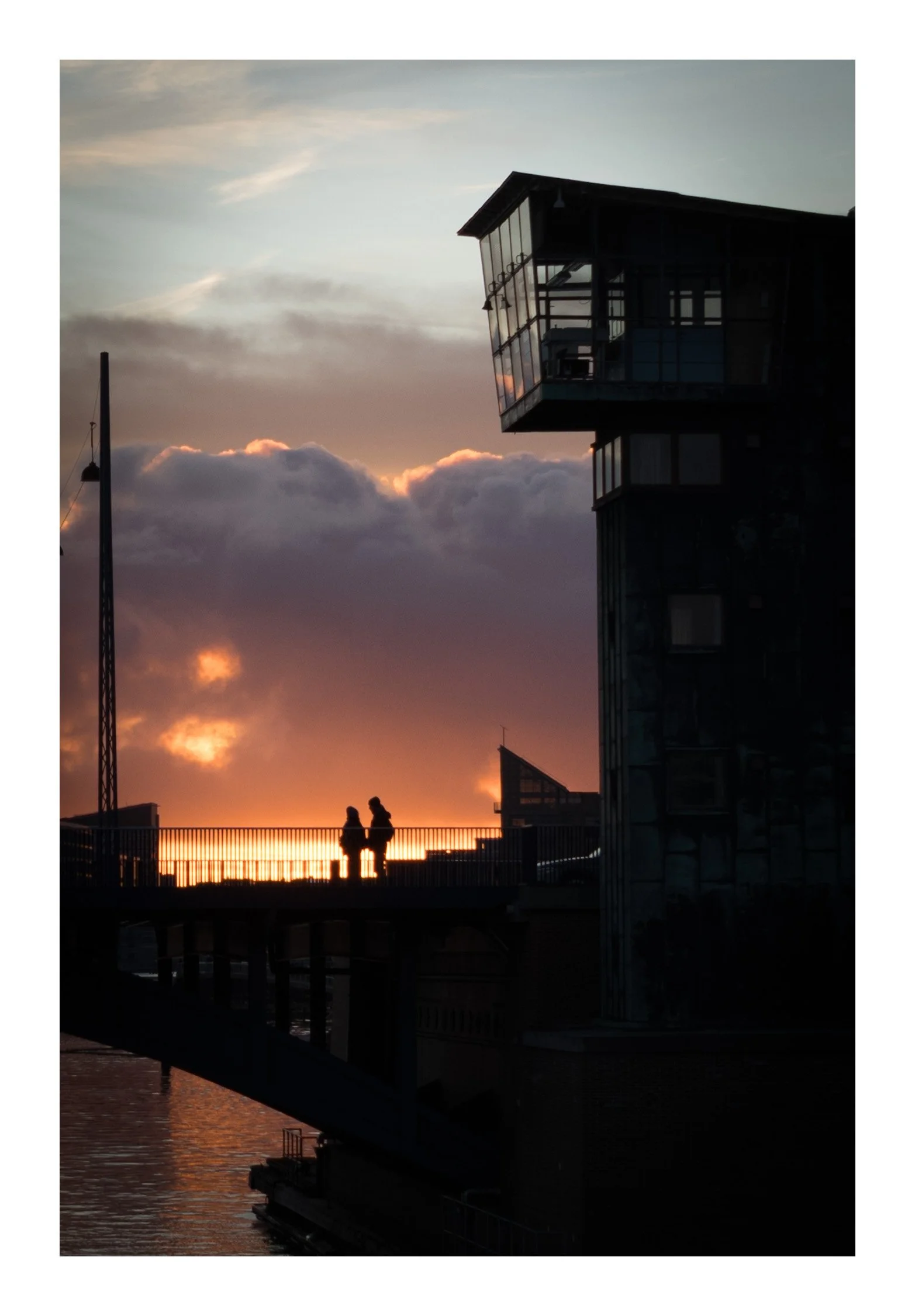 A fiery sunset glows behind the clouds as the sky turns deep orange and red. On the bridge, two people stand quietly, with the silhouette of the bridge tower rising beside them. It feels calm, dramatic, and a little bit magical — the perfect end to t