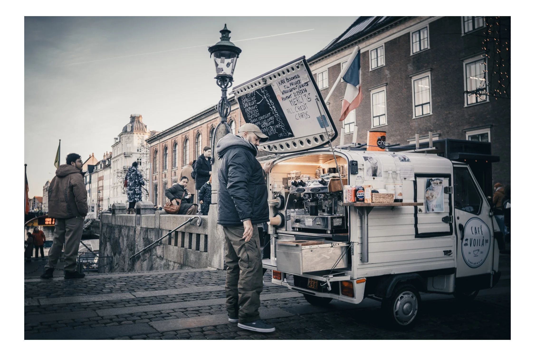 In a tiny coffee van, someone stands at the espresso machine, grinding beans and steaming milk while the window opens to the street. The air smells like fresh coffee, and people stop by for a warm cup and a quick smile.