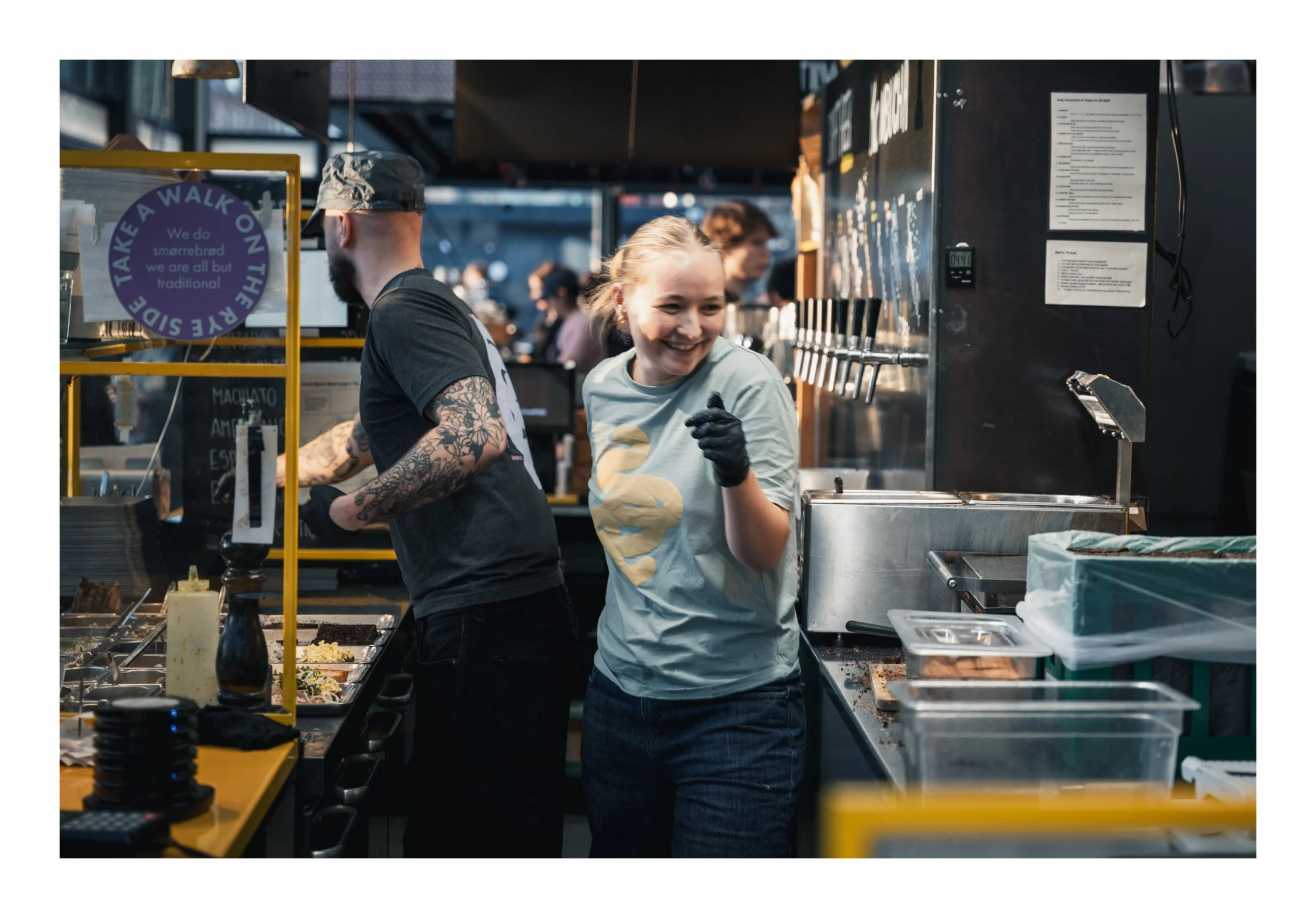 A woman stands behind the counter carefully making a smørrebrød, spreading butter, layering fresh toppings, and finishing it with love. As she works, she smiles — calm, warm, and genuinely happy — like she’s sharing a little piece of Danish tradition