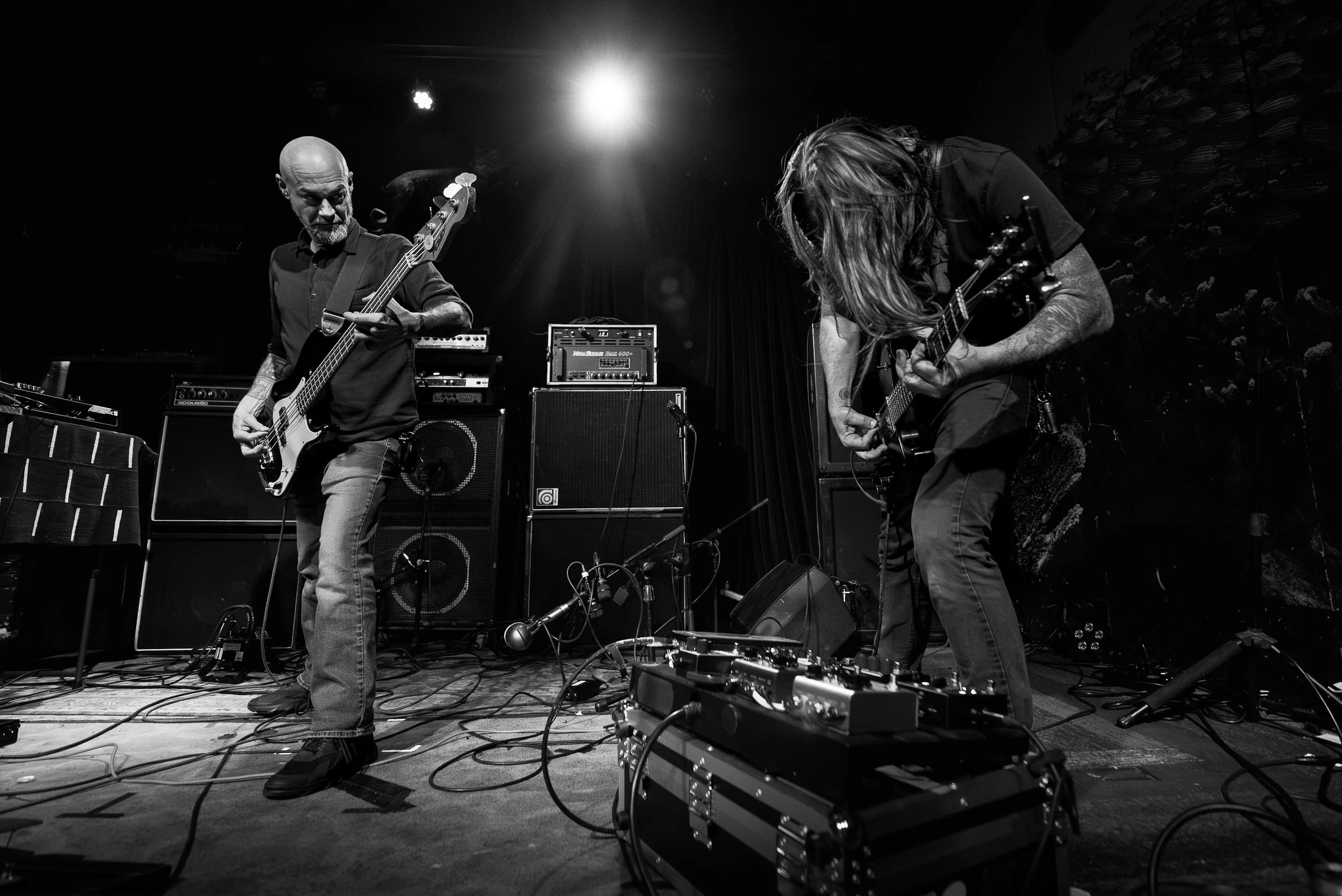 Two musicians performing on stage, one playing bass guitar and the other playing electric guitar, with amplifiers and equipment around them, under stage lights.
