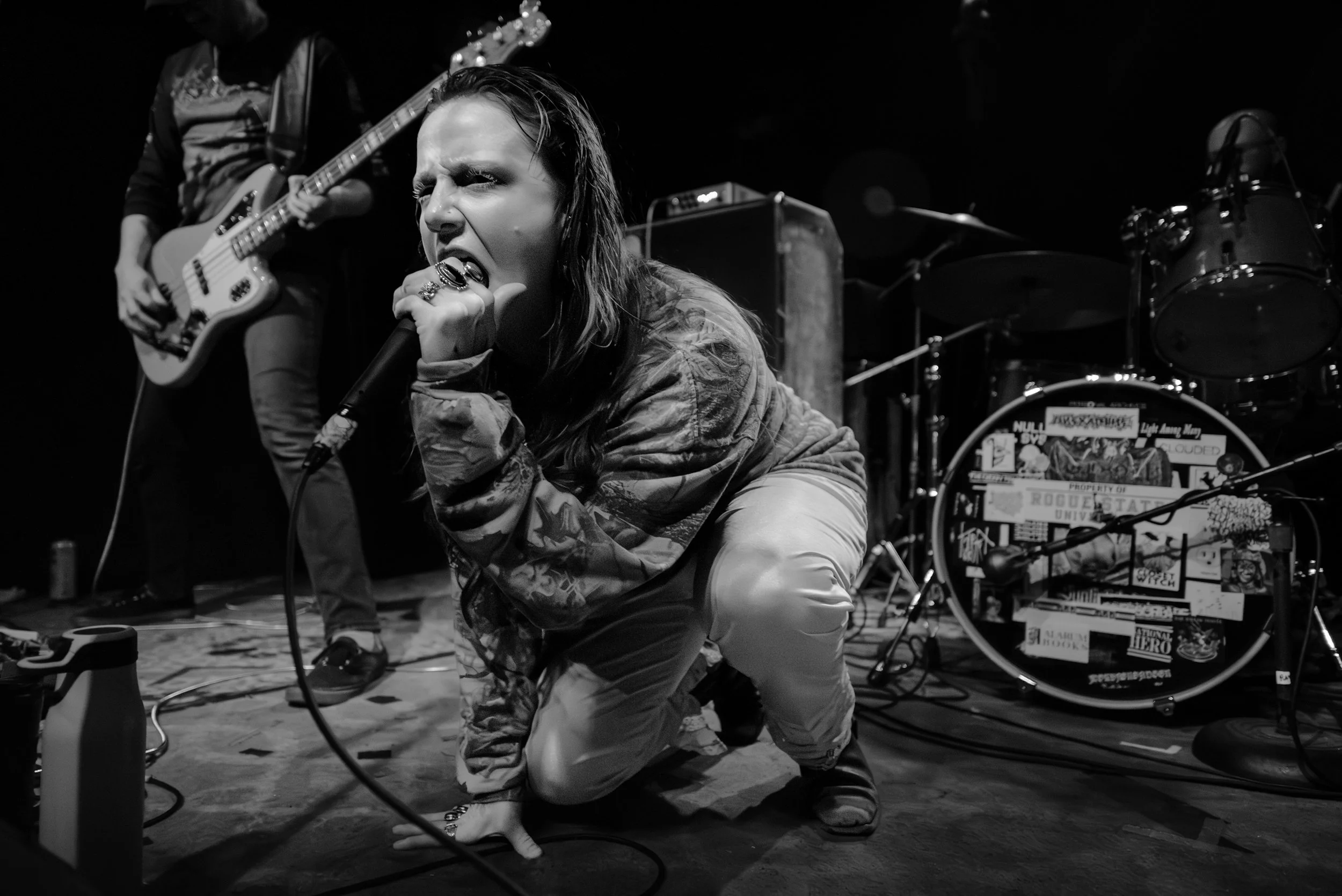 A female vocalist passionately singing into a microphone on stage, with a guitarist in the background and a drum set covered in stickers behind her, in a black and white photo.