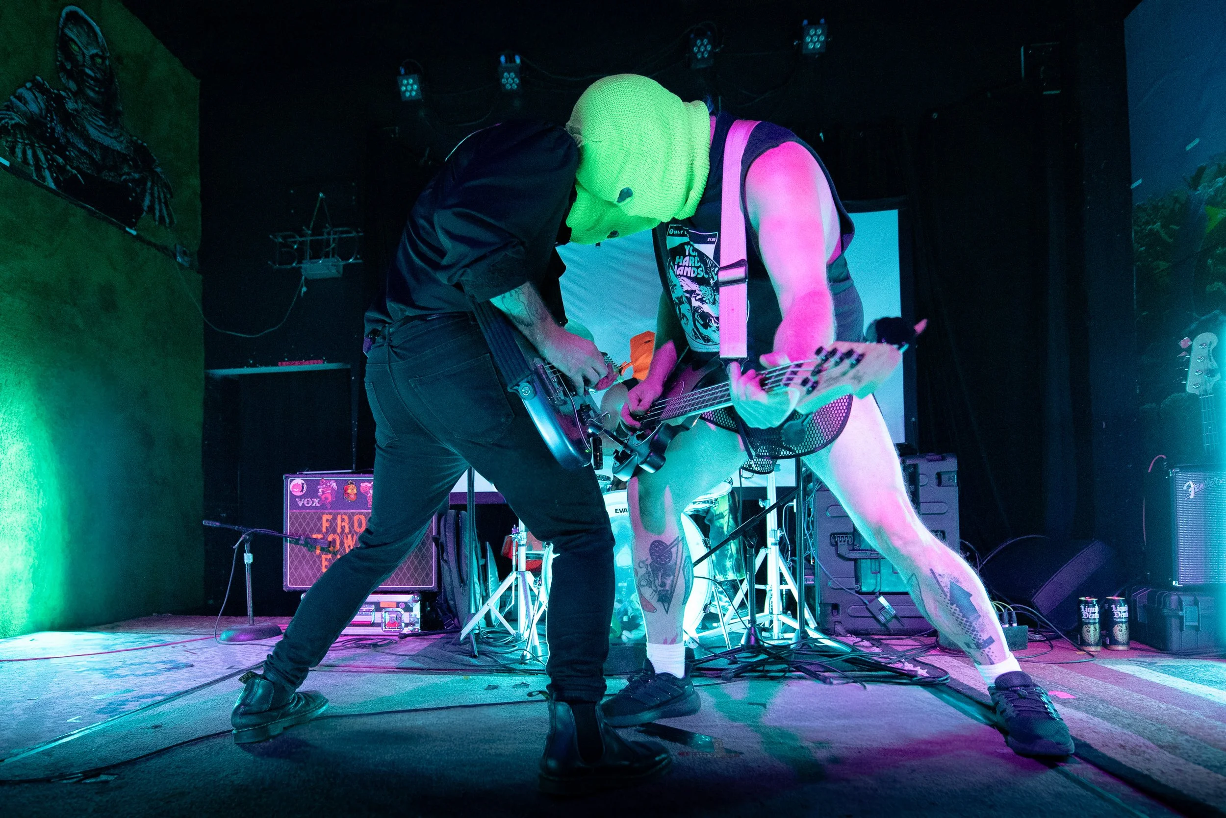 Two musicians wearing neon green masks and colorful clothing perform on stage with guitars, standing close together under concert lights.