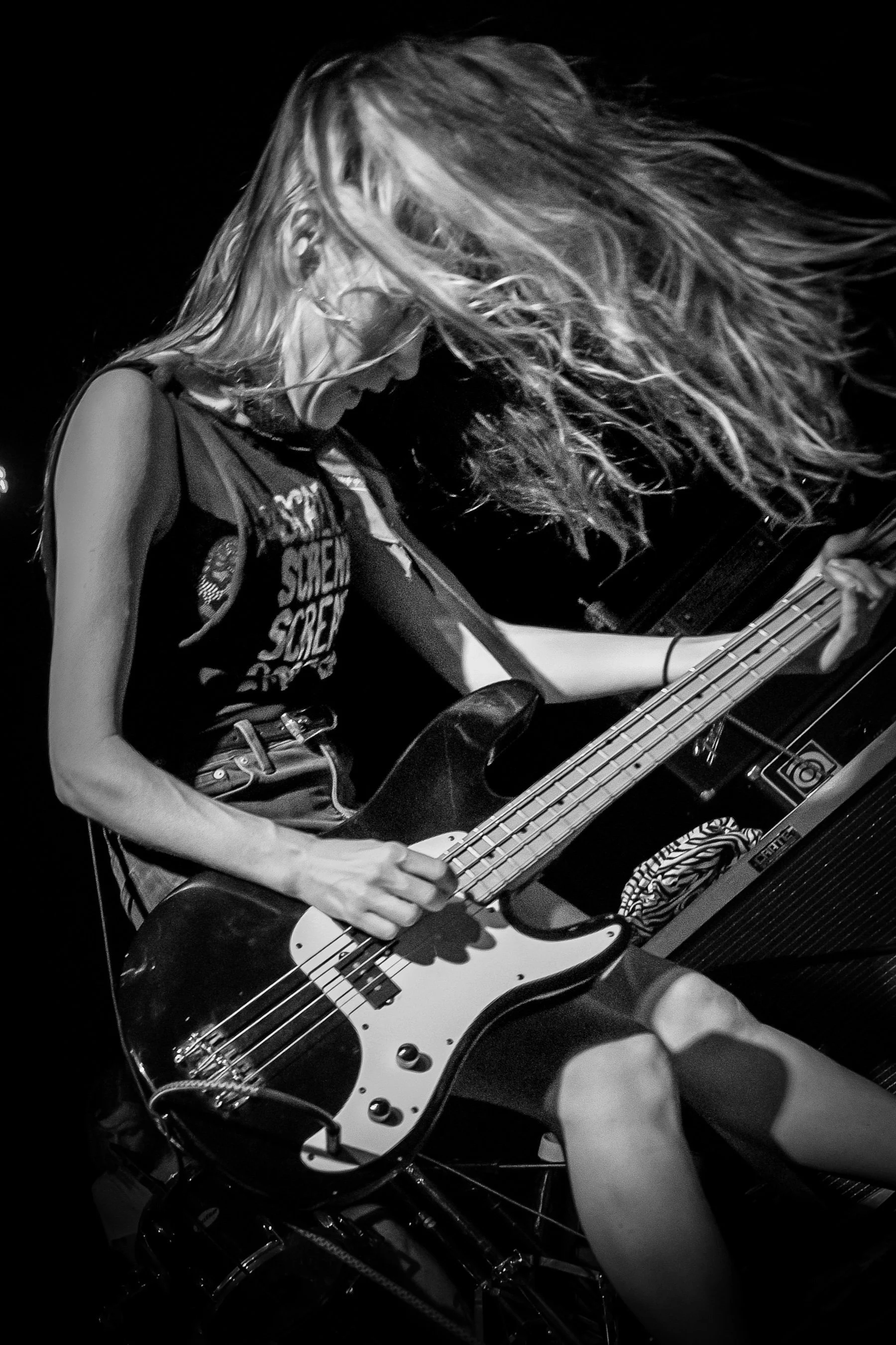 A female musician playing a bass guitar during a concert, with long hair flowing and wearing a sleeveless shirt.
