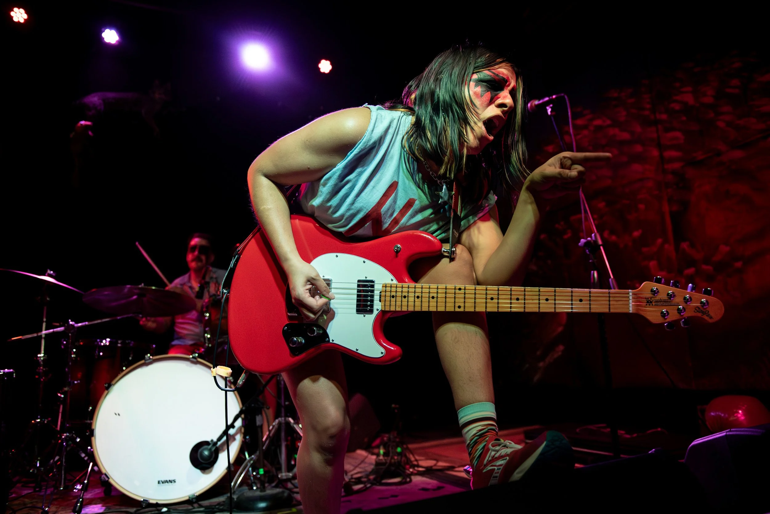 A female musician with colorful face paint, wearing a sleeveless top and knee-high socks, passionately performs on stage with a red electric guitar. A drummer is visible in the background behind a drum set, under stage lights.