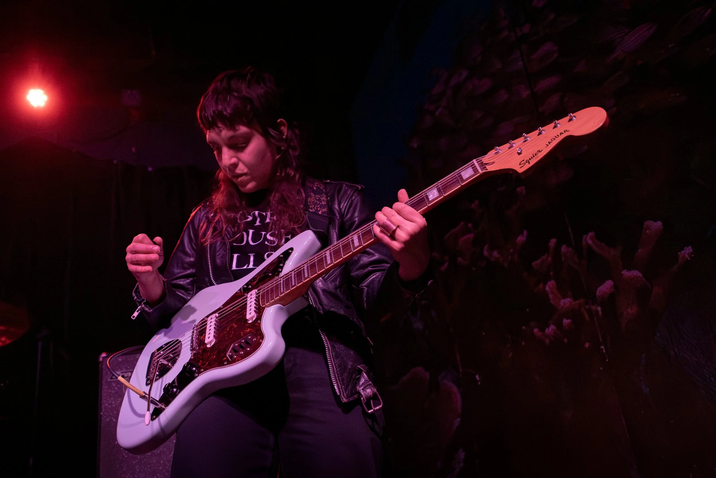 Female musician playing an electric guitar on stage under red and purple lighting.