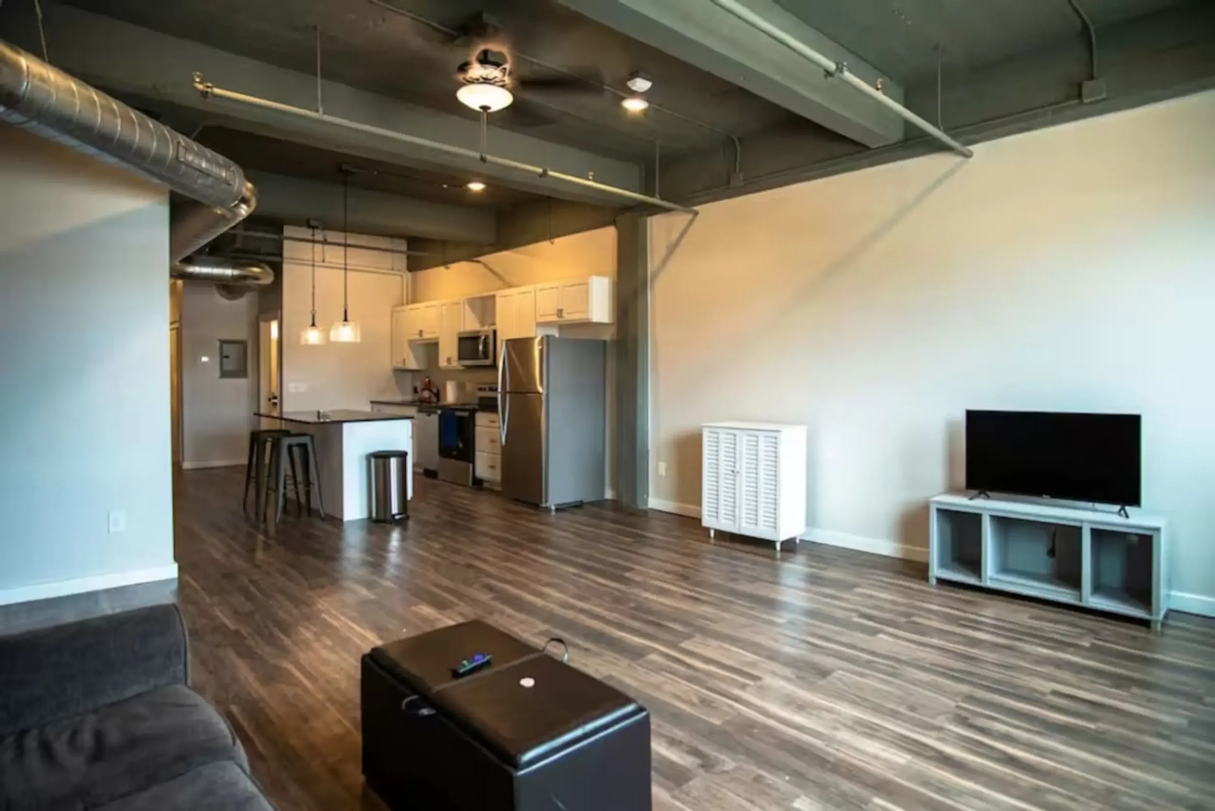 Open-concept living room and kitchen area with hardwood floors, a small TV on a silver stand, a white radiator cover, and kitchen appliances including a microwave and refrigerator. The space features exposed ductwork and a concrete ceiling with ceiling fans and lighting.