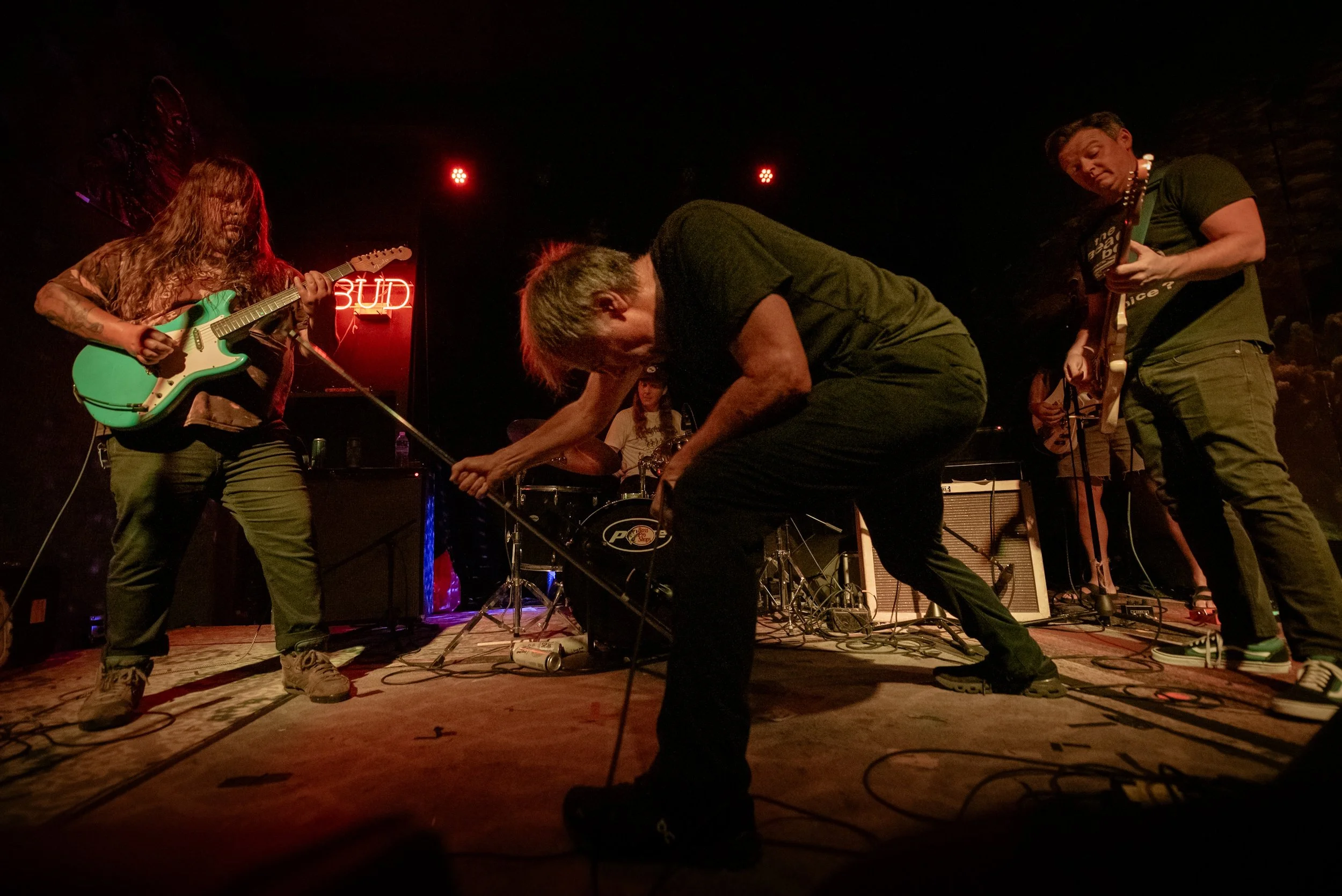 A band performing live on stage with musicians playing guitars and drums, under red lighting, in a dark venue.