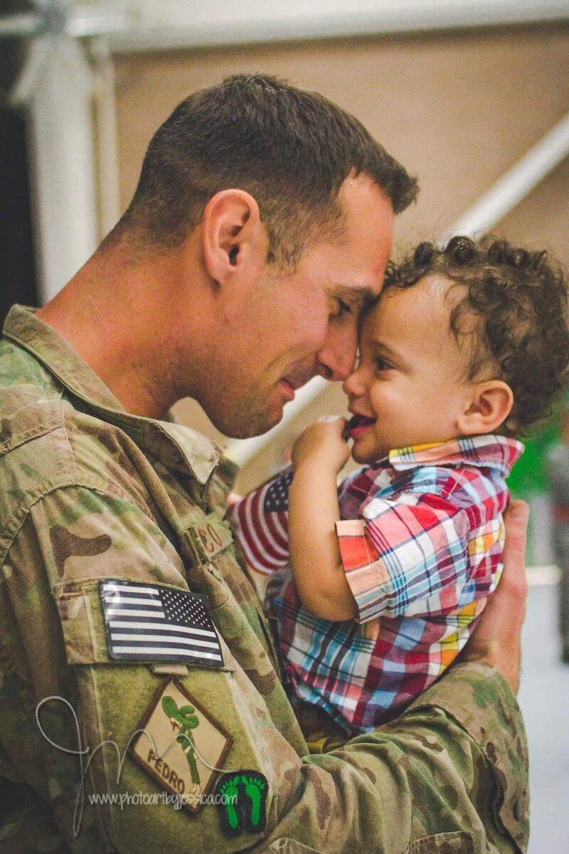 A soldier in camouflage uniform holding a smiling young child with curly hair close to his face, touching foreheads, indoors.