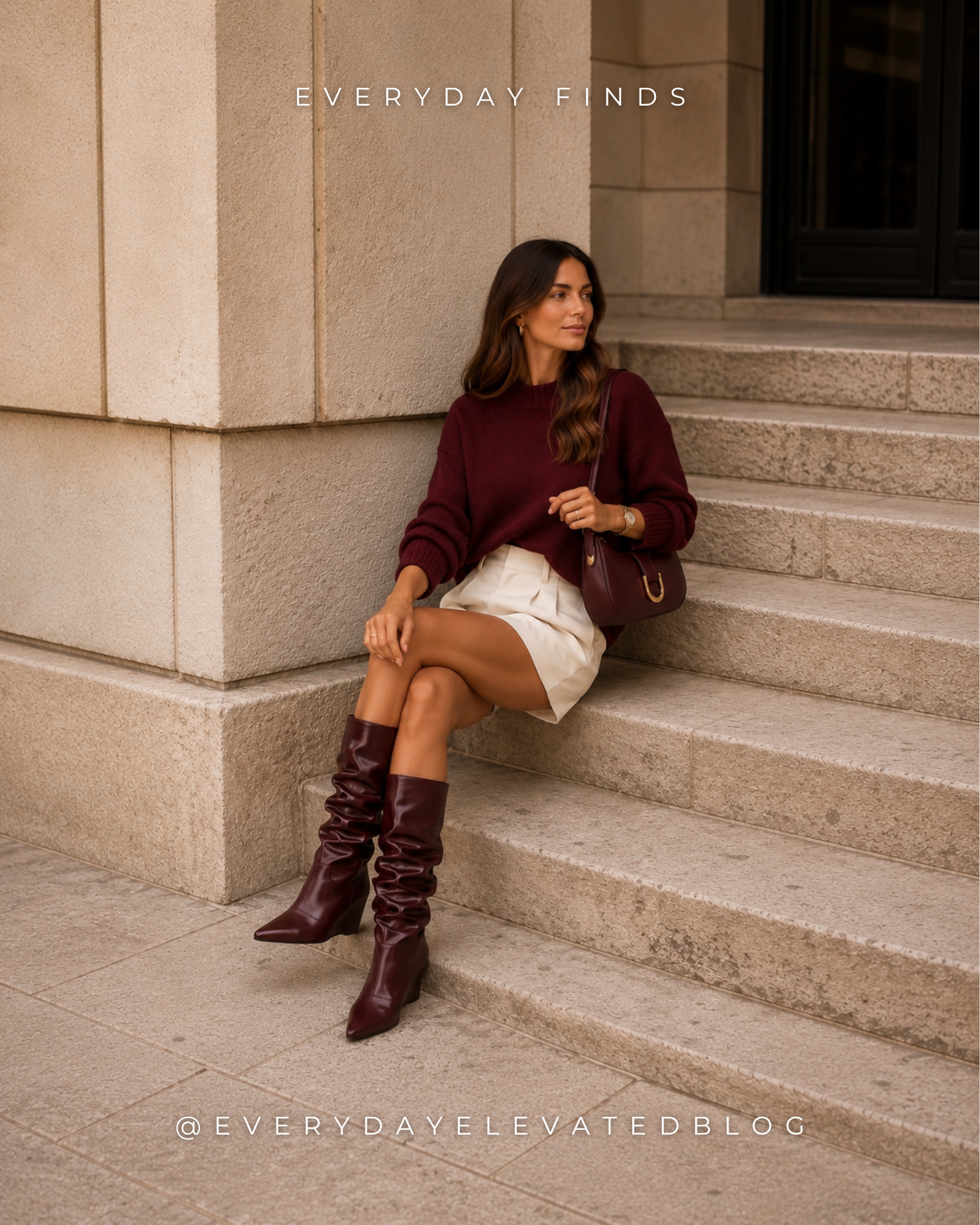 Woman wearing a burgundy sweater, cream tailored shorts, burgundy slouchy knee-high boots, and a matching burgundy shoulder bag while walking through an upscale city setting, showcasing an elevated fall shorts outfit for warm early fall weather.