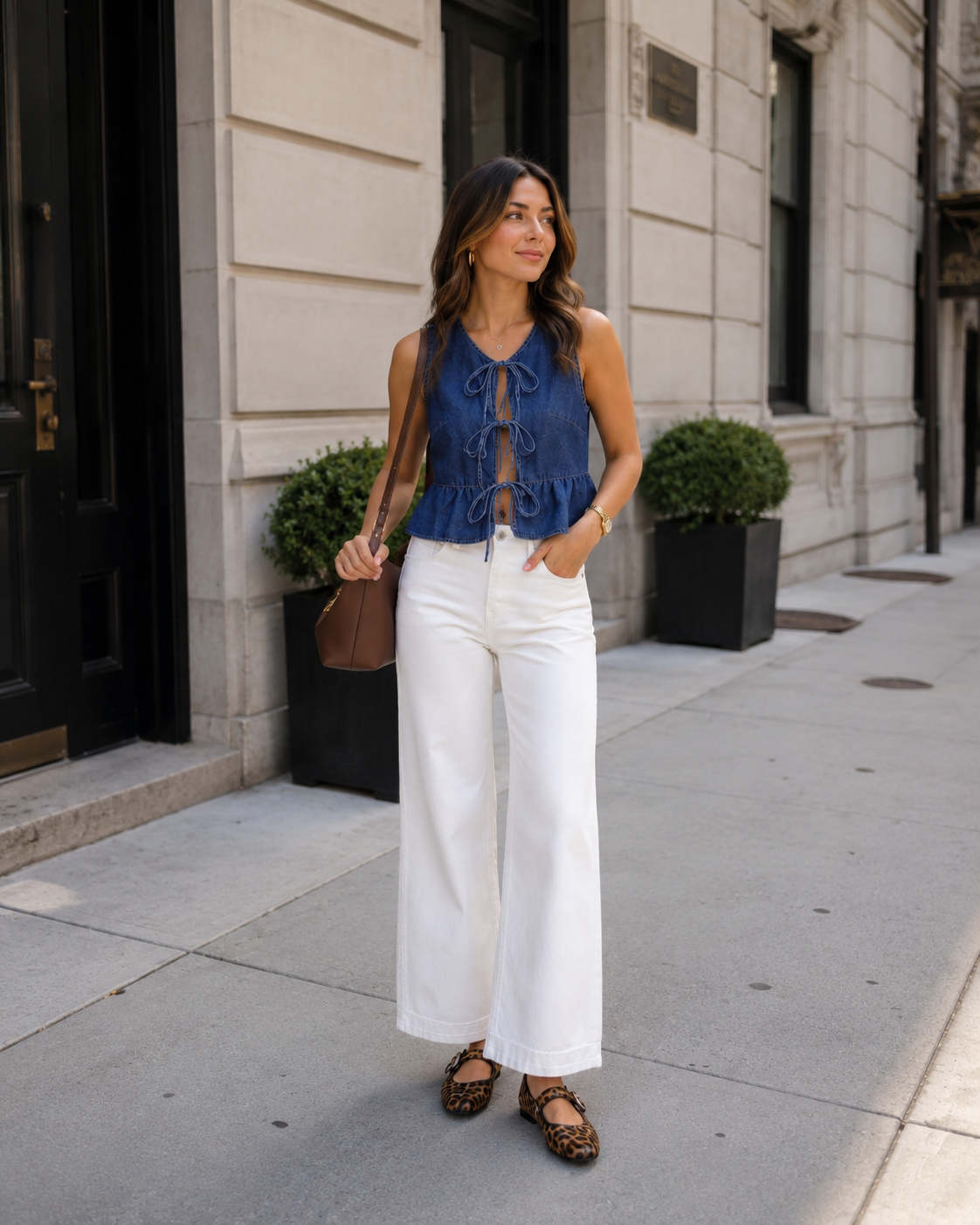 Woman wearing a casual spring city outfit with a denim tie-front peplum top, white wide leg jeans, leopard print Mary Jane flats, and a brown shoulder bag while walking on a clean upscale city street with neutral architecture and gold accessories.