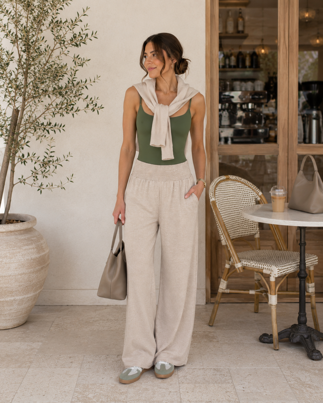 Woman wearing an olive green bodysuit, beige wide-leg pants, sneakers, and a lightweight sweater draped over her shoulders in a casual chic summer outfit perfect for errands and everyday wear.